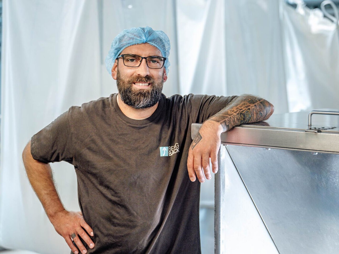 Bearded warehouse worker in hairnet and glasses smiling by industrial stainless conveyor in food processing facility