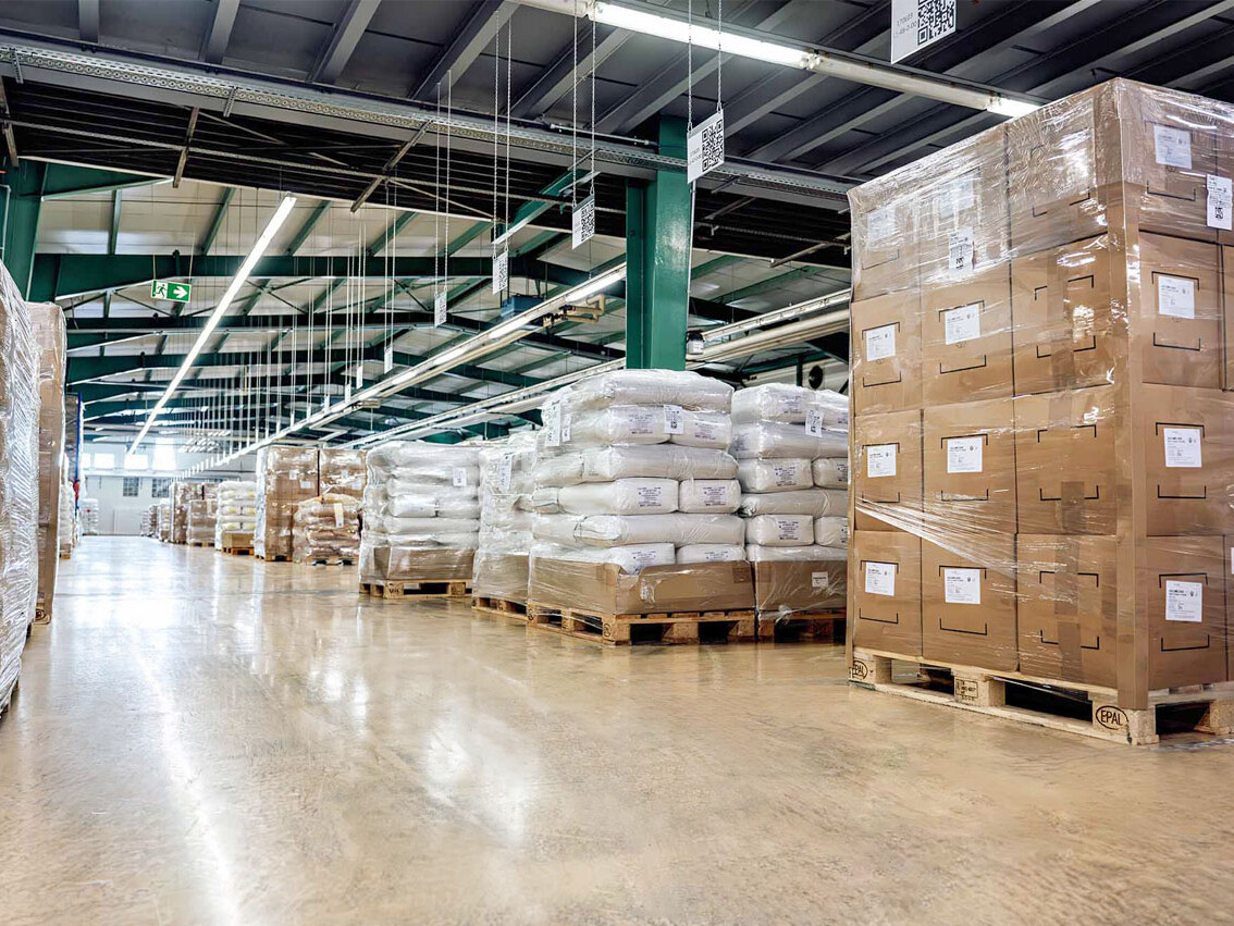 Warehouse interior with stacked pallets of packaged goods, plastic-wrapped bundles, and large cardboard boxes on pallets