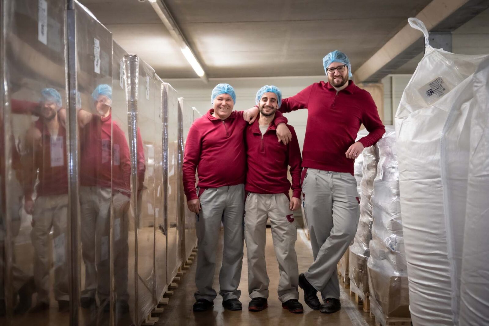 Three workers in red jackets wearing hairnets and gloves standing in a warehouse hallway among packaged supplies