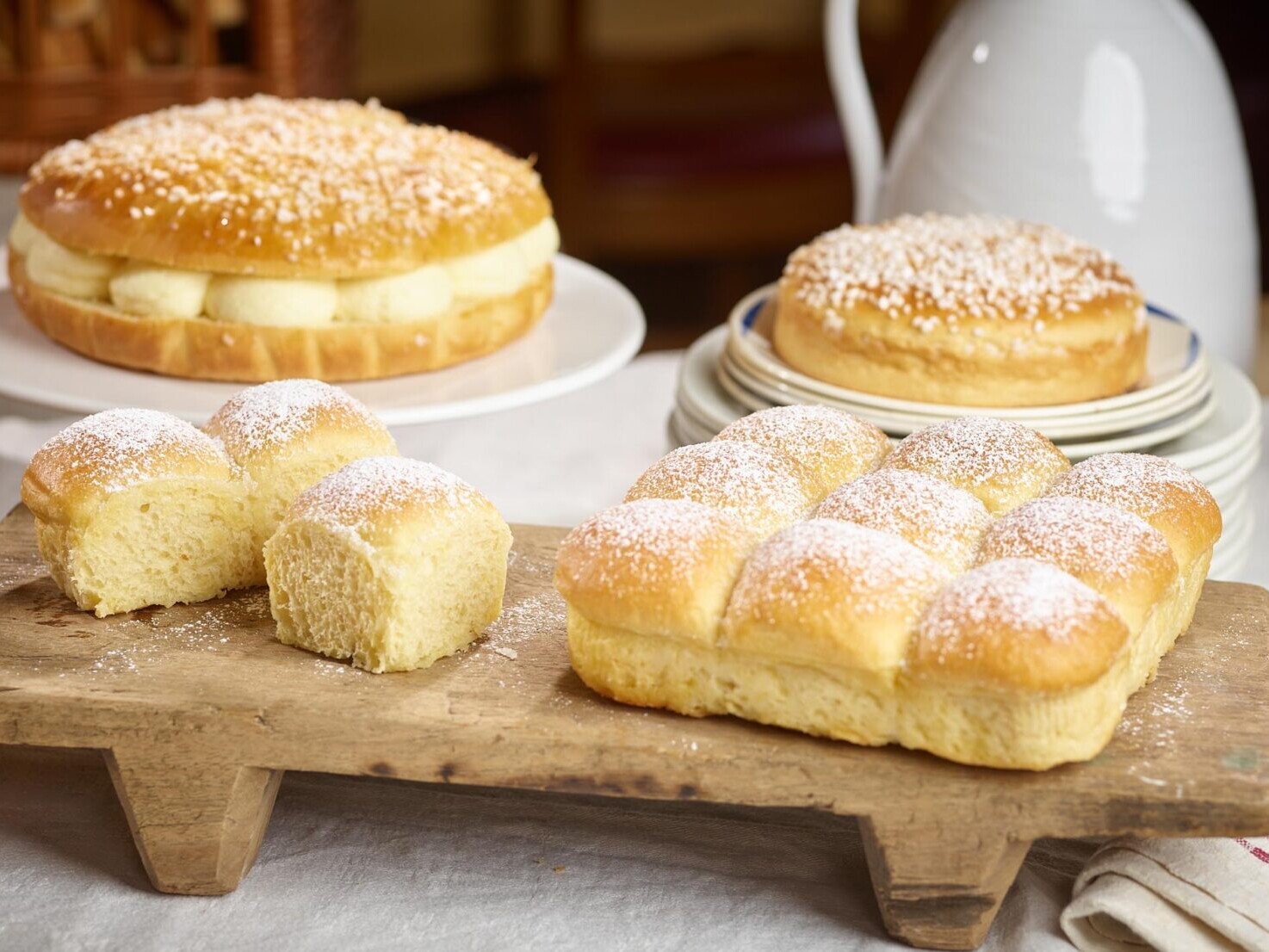 Fresh baked brioche rolls and loaf on a rustic wooden board dusted with sugar, with pastries in background