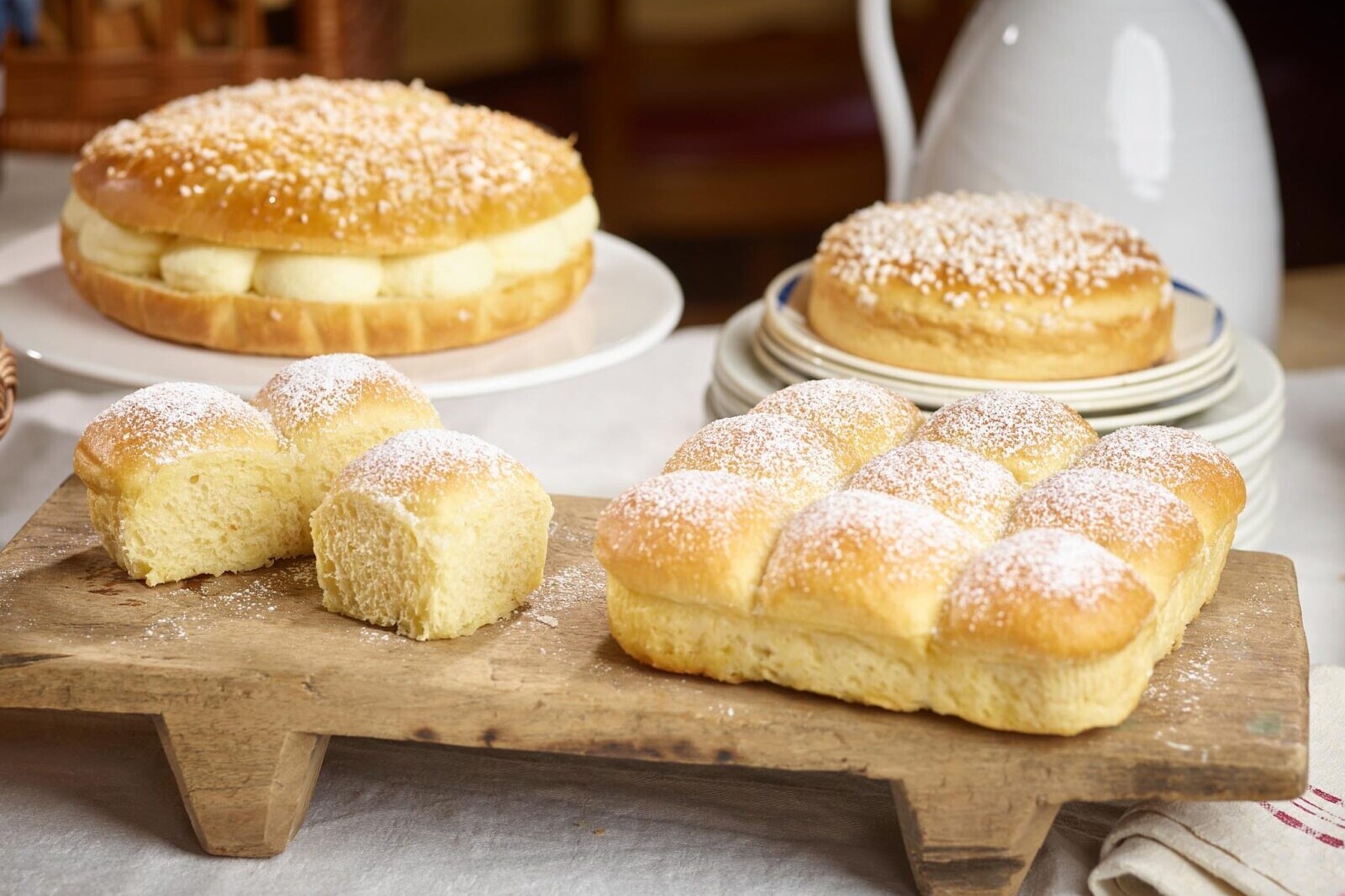 Fresh baked brioche rolls and loaf on a rustic wooden board dusted with sugar, with pastries in background