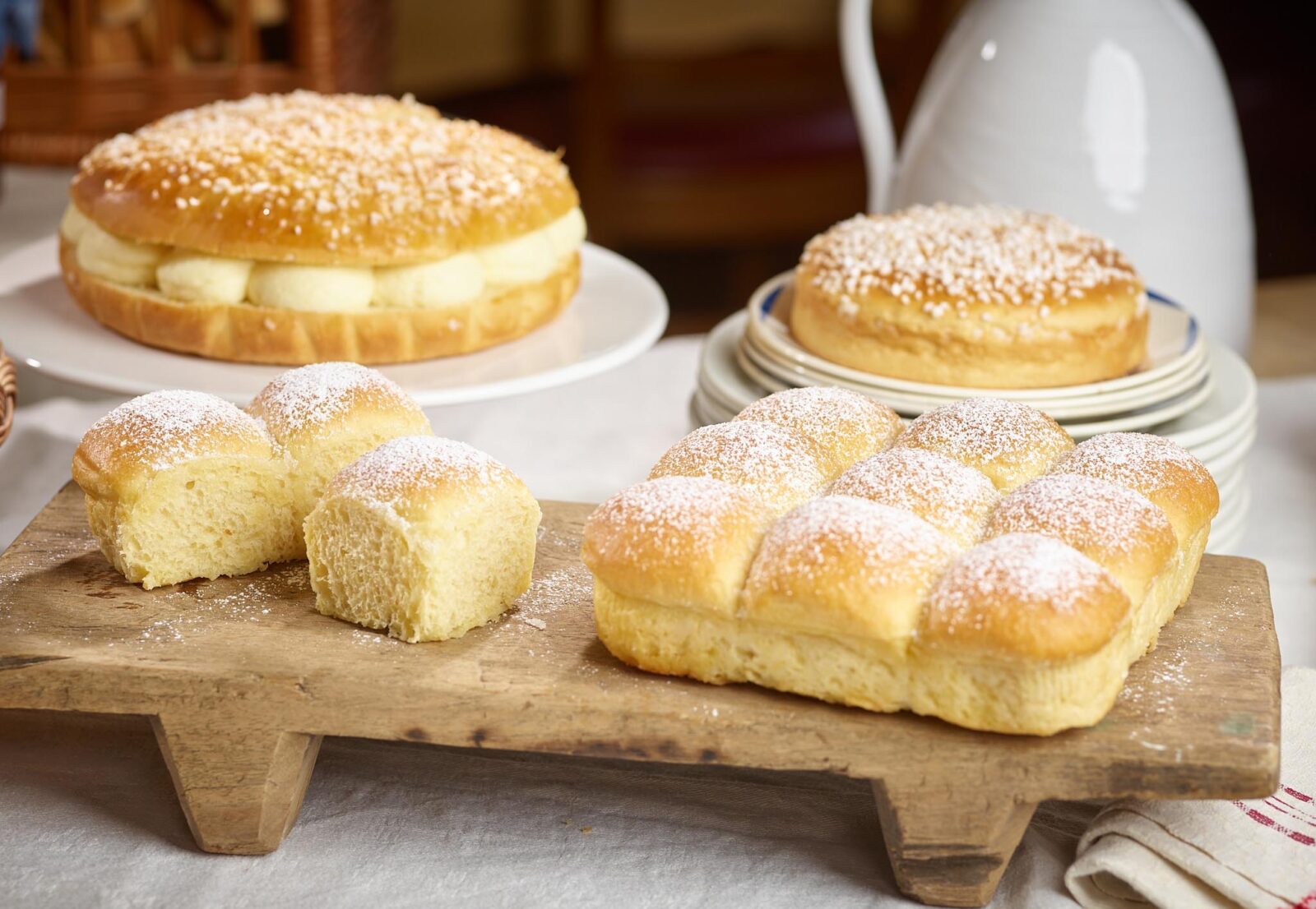 Fresh baked brioche rolls and loaf on a rustic wooden board dusted with sugar, with pastries in background