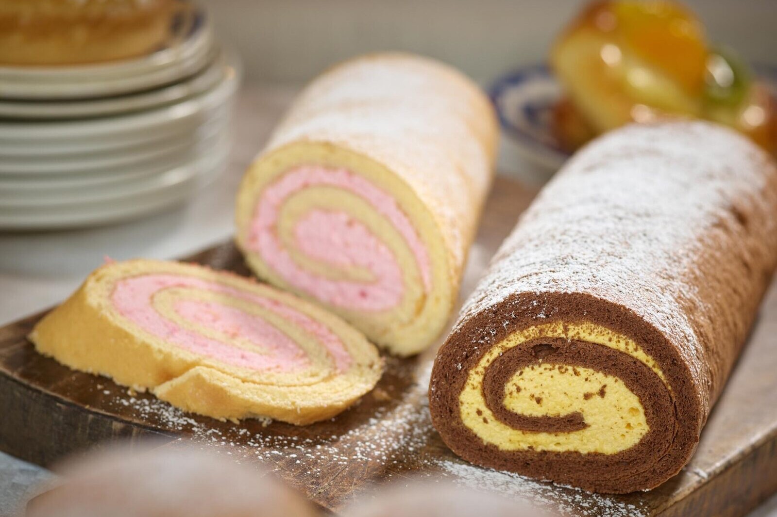 Pinwheel cake rolls with pink and yellow swirl slices on a wooden board dusted with powdered sugar