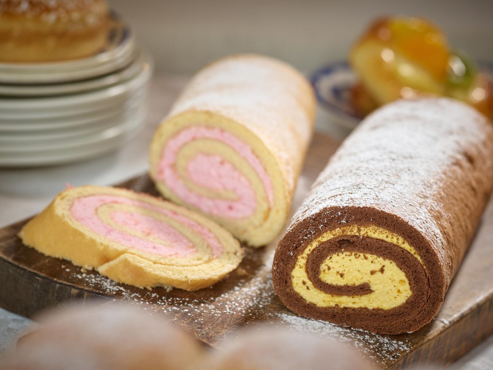 Pinwheel cake rolls with pink and yellow swirl slices on a wooden board dusted with powdered sugar