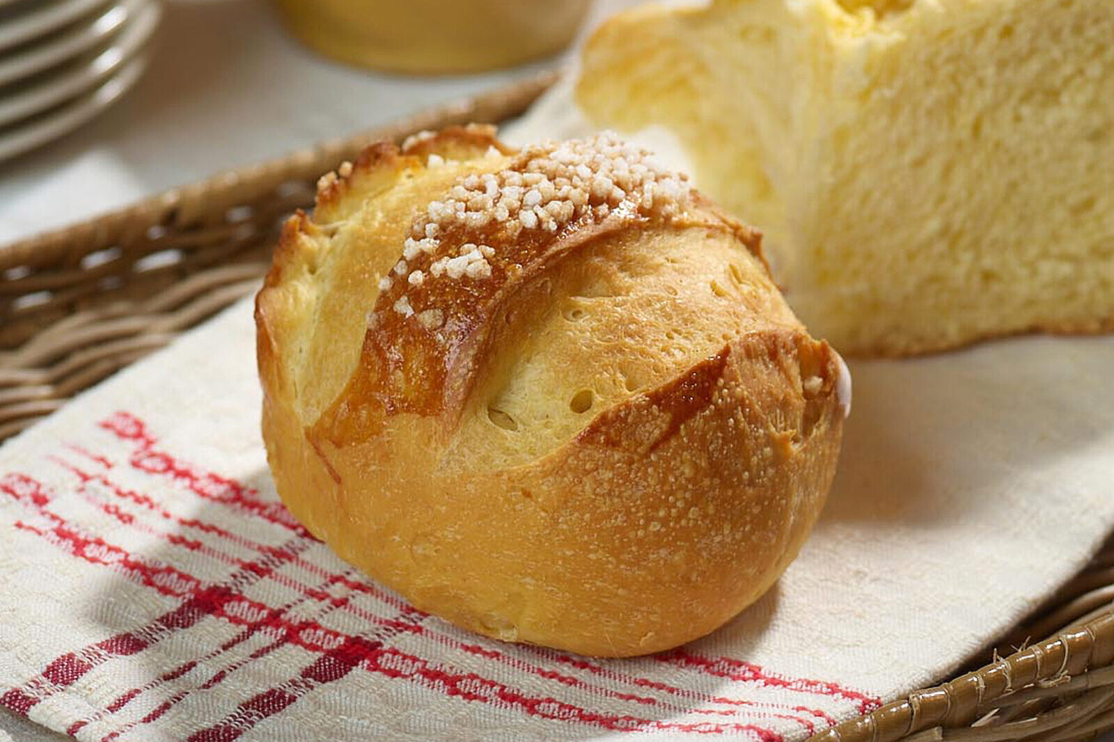Rustic baked bread roll with golden crust and coarse salt on a striped cloth, with basket and loaf in background