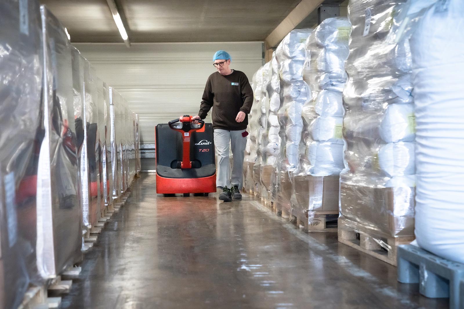 Warehouse worker operating a Linde T20 pallet jack, moving through an aisle packed with stored goods