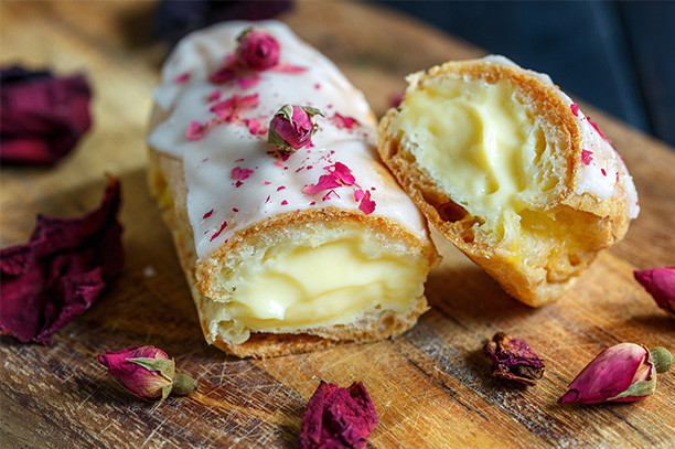 Cream-filled vanilla éclairs with rose petals on rustic wooden board, close-up dessert photo