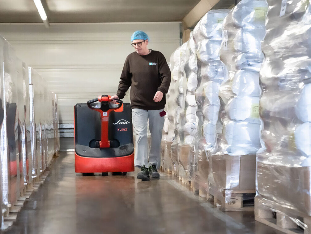 Warehouse worker operating a Linde T20 pallet truck, moving through plastic-wrapped storage aisles.