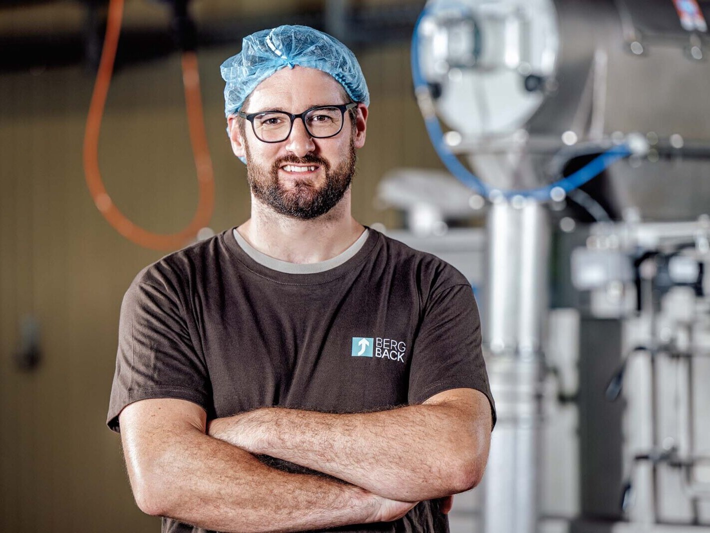 Food factory worker wearing hair net and glasses, standing with arms crossed near industrial machinery