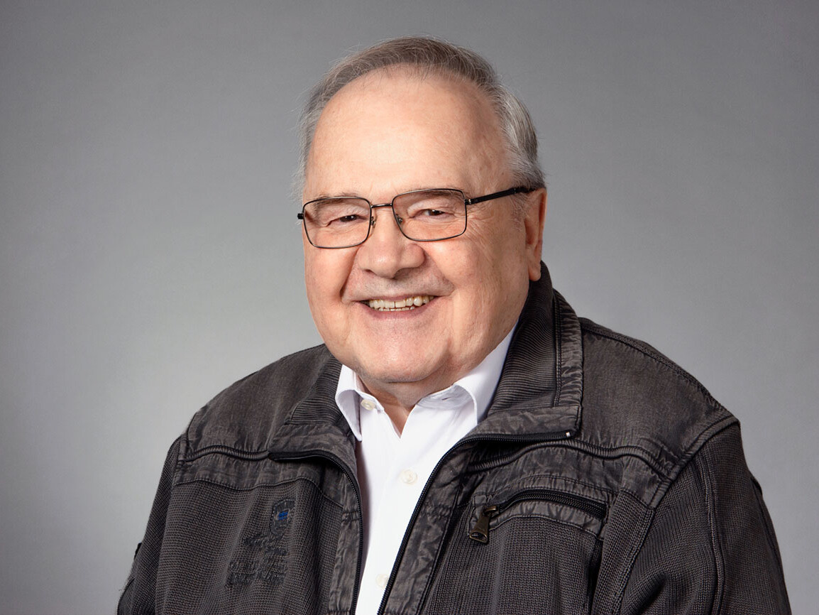 Smiling elderly man wearing glasses and a gray jacket against a neutral background.