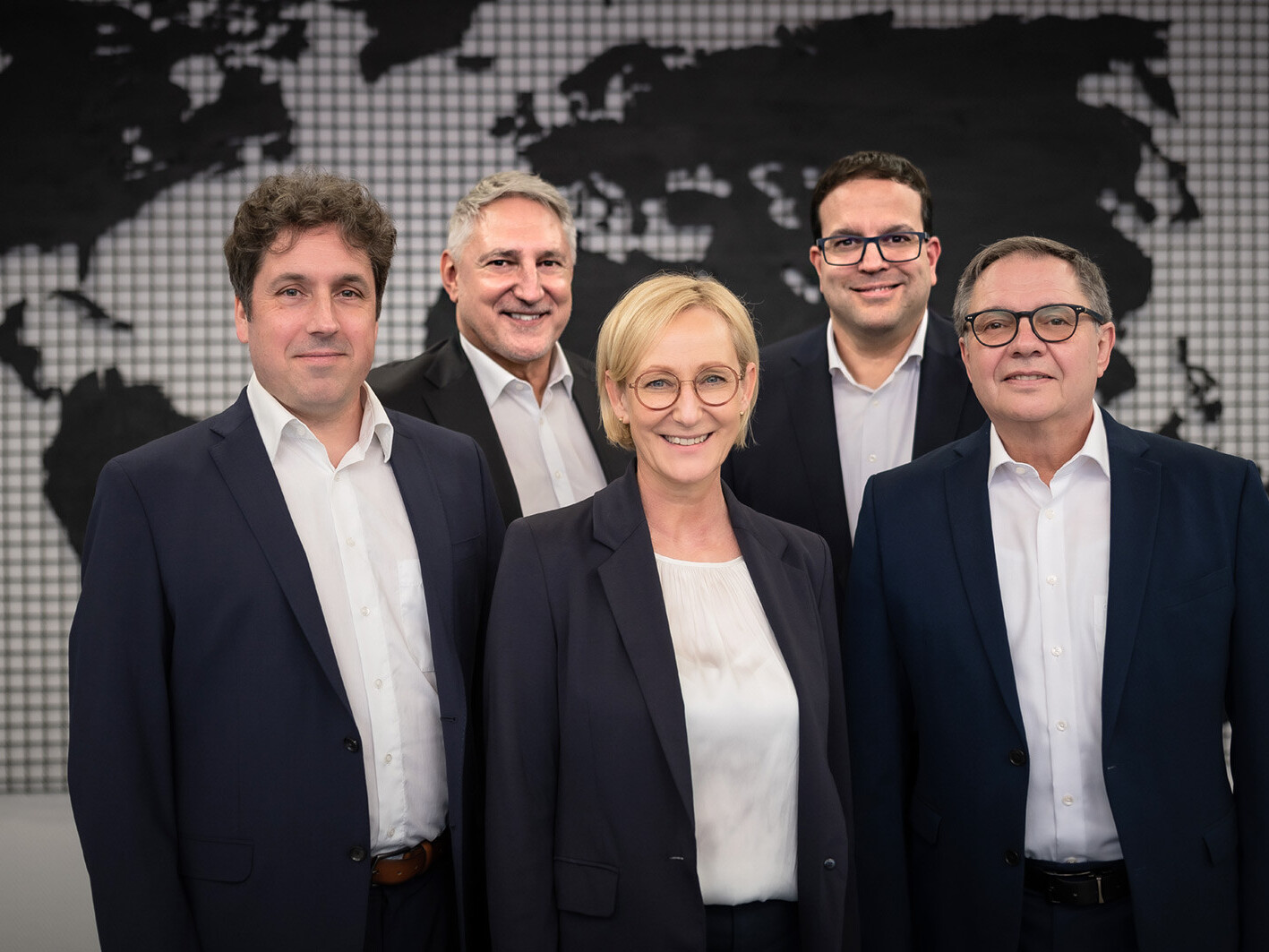 Five business professionals in suits standing in front of a world map background, smiling.